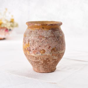 May include: A small, antique, weathered terracotta pot with a rounded body and a slightly flared rim. The pot has a rough, textured surface with shades of brown, orange, and yellow. It appears to be aged, with visible wear and tear.