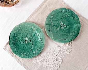 Puede incluir: Dos platos redondos de cerámica con un esmalte verde intenso, cada uno con un patrón en relieve detallado de hojas superpuestas en el centro y un diseño de rejilla texturizada a lo largo de los bordes festoneados, creando un aspecto botánico.