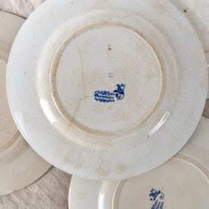 May include: A collection of antique white ceramic plates with visible signs of age and wear. The plates have a faded, crackled glaze and a blue floral emblem on the underside. The plates are arranged in a slightly overlapping pattern.