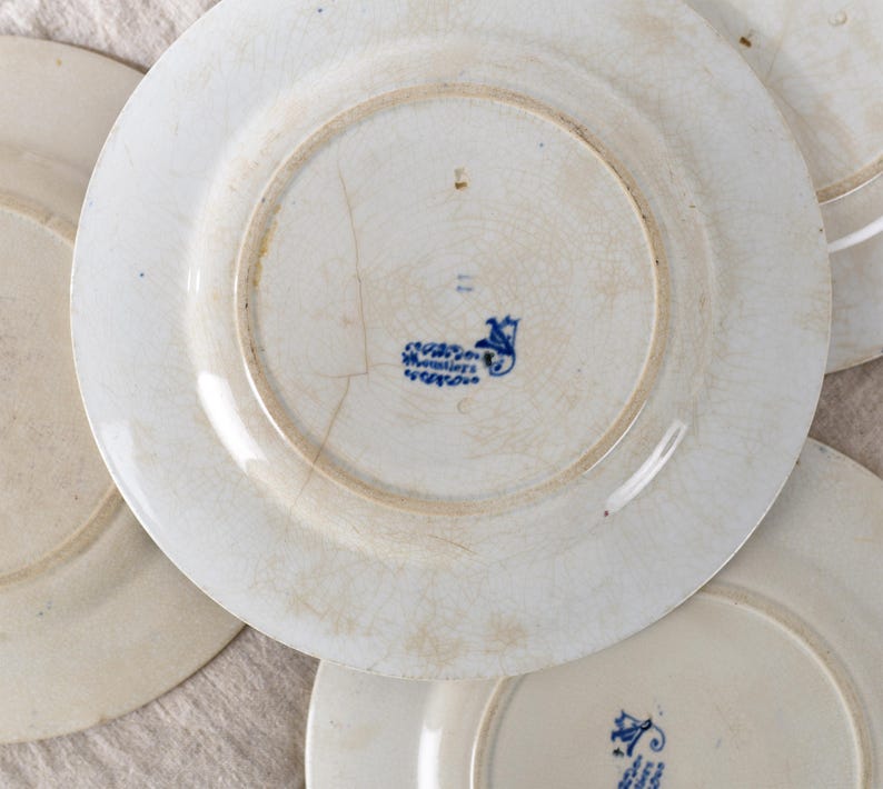 May include: A collection of antique white ceramic plates with visible signs of age and wear. The plates have a faded, crackled glaze and a blue floral emblem on the underside. The plates are arranged in a slightly overlapping pattern.