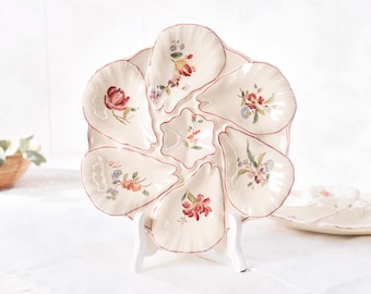 May include: A cream-colored, multi-sectioned serving platter with a floral design. Each section has a scalloped edge with a red trim. The platter is supported by a white stand. The floral designs include red, pink, and yellow flowers.