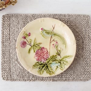 Könnte beinhalten: Ein runder, cremefarbener Keramikteller mit einem erhabenen, dreidimensionalen Reliefdesign. Das Dekor zeigt einen grünen Vogel auf einem Zweig, umgeben von detaillierten rosa Blüten und grünen Blättern, mit einem gewellten, geprägten Rand.