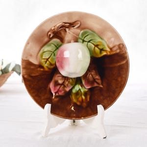 Könnte beinhalten: Ein dekorativer Keramikteller mit einem erhabenen Apfel- und Blattdesign. Der Teller hat einen braunen Hintergrund mit grünen, rosa und gelben Blättern. Ein weißer und rosa Apfel befindet sich in der Mitte. Der Teller steht auf einem weißen Ständer.