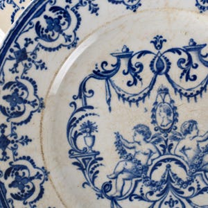 May include: Close-up of a white and blue porcelain plate. The plate features a detailed blue floral and cherub design. The rim has a blue border with intricate patterns. The plate's design includes cherubs, floral elements, and a portrait.