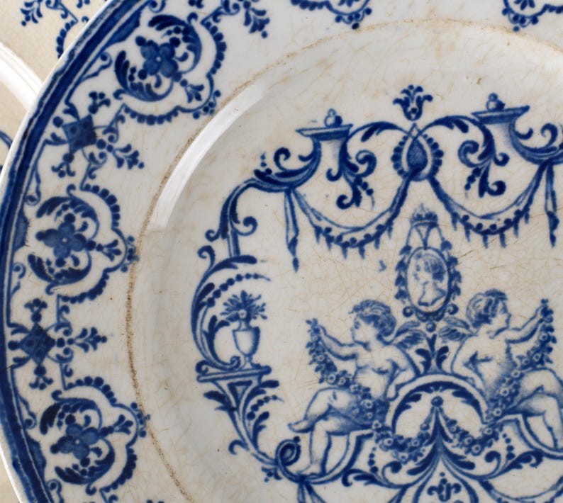 May include: Close-up of a white and blue porcelain plate. The plate features a detailed blue floral and cherub design. The rim has a blue border with intricate patterns. The plate's design includes cherubs, floral elements, and a portrait.