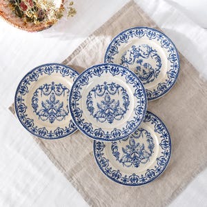 May include: Four cream-colored plates with blue floral and ornate designs. Each plate has a blue border and a central motif of figures and decorative elements. The plates are arranged on a neutral-toned linen cloth.