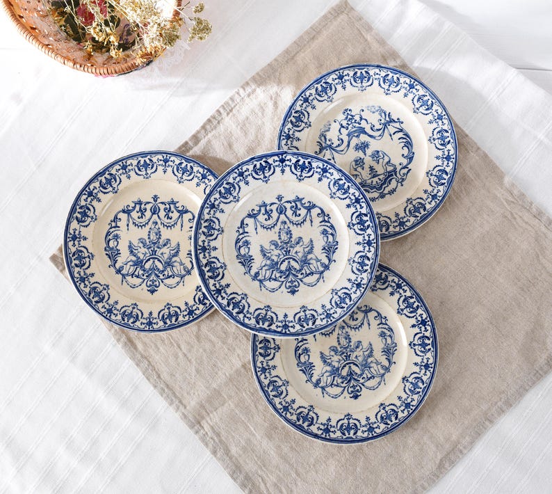 May include: Four cream-colored plates with blue floral and ornate designs. Each plate has a blue border and a central motif of figures and decorative elements. The plates are arranged on a neutral-toned linen cloth.