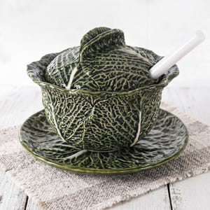 Peut inclure: Un bol en céramique vert foncé avec un couvercle et une soucoupe assortis, tous dotés d'une texture en relief représentant des feuilles de chou. Le bol comprend une cuillère en céramique blanche insérée dans une ouverture du couvercle.