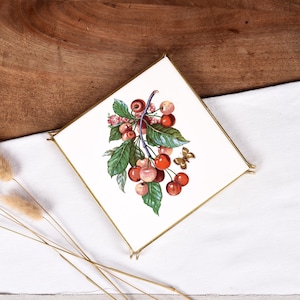 Puede incluir: Un posavasos de cerámica blanca con marco de metal dorado presenta una ilustración vintage de una rama de cerezo con cerezas rojas y rosadas y una mariposa.
