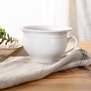 May include: White ceramic chamber pot with a rounded body and a single handle. The pot is sitting on a light brown linen cloth with black stripes.