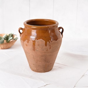 Puede incluir: Una gran olla de terracota antigua con una abertura ancha y dos asas pequeñas. La olla tiene una parte superior vidriada marrón brillante que gotea por los lados, contrastando con la parte inferior rugosa y sin vidriar. La olla está sobre una superficie blanca.