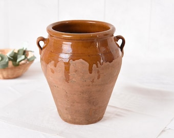 May include: A large, antique terracotta pot with a wide opening and two small handles. The pot has a glossy, brown glazed top that drips down the sides, contrasting with the rough, unglazed lower portion. The pot sits on a white surface.