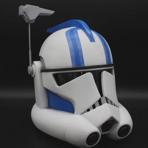 Puede incluir: Un casco blanco y azul con visera y antena gris. El casco tiene rayas azules en la parte superior y detalles azules en los laterales. El casco es una réplica de un casco de soldado clon de Star Wars.