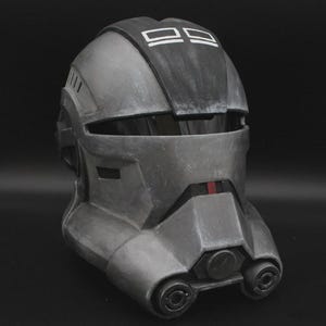 May include: A gray and black helmet with a weathered appearance. The helmet has a visor opening and features a black top section with two white rectangles. The front has a red accent and two circular details.