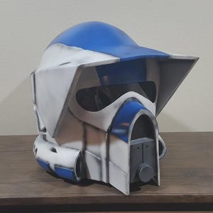 May include: A blue and white helmet with a visor, likely a costume piece. The helmet has a rounded top and angular features, with a dark visor area. The color scheme is primarily white and blue, with some gray accents. The helmet is sitting on a wooden surface.