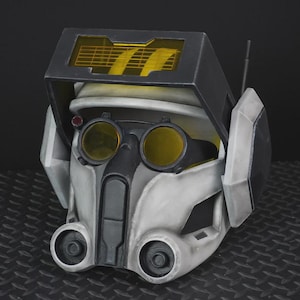 May include: A detailed, futuristic helmet in shades of gray, black, and yellow. The helmet features a visor with yellow lenses, a rectangular top section, and circular details on the lower front. A small antenna extends from the side.