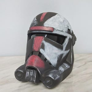 May include: A black and gray helmet with red and white accents. The helmet has a visor and is painted with a distressed finish. The design is reminiscent of a military or science fiction theme.