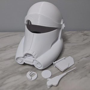 May include: A white, 3D-printed helmet with a distinctive design, reminiscent of a stormtrooper helmet. The helmet is accompanied by several detached components, including a visor, a circular piece, and a long, thin piece. The items are on a marble-patterned surface.
