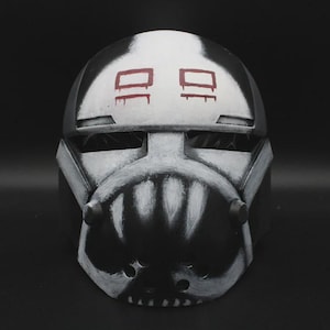May include: A black and white helmet with red rectangular markings. The helmet has a stylized design with a face-like appearance, featuring a mouth-like opening and eye-like shapes. The overall aesthetic is reminiscent of a futuristic or sci-fi theme.