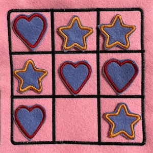 May include: A pink felt board with a black grid pattern. The grid has nine squares, each containing a felt heart or star. The hearts are blue with red outlines, and the stars are blue with yellow outlines.