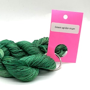 May include: A twisted skein of green embroidery thread featuring varying shades of emerald and forest green. The thread is attached to a bright pink rectangular paper tag with a white label that reads Green spider mum and a silver metal ring.
