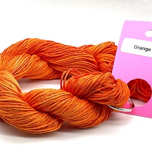 Puede incluir: Una madeja de hilo de bordar de color naranja vibrante retorcida en un bucle y unida a una etiqueta de papel rectangular de color rosa brillante. La etiqueta tiene un rótulo blanco con el texto Orange crush y un orificio circular.