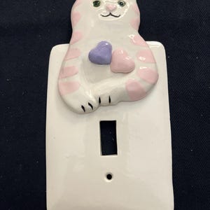 May include: A white ceramic light switch cover featuring a cat design. The cat has pink spots and holds two hearts, one purple and one pink. The cat has green eyes and a black nose and whiskers.