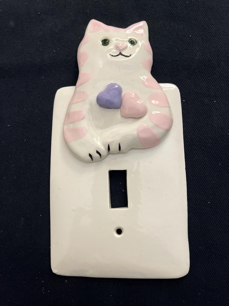 May include: A white ceramic light switch cover featuring a cat design. The cat has pink spots and holds two hearts, one purple and one pink. The cat has green eyes and a black nose and whiskers.