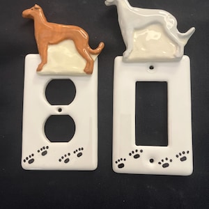 May include: Two white ceramic light switch plates featuring raised dog figures. One plate has a brown dog and a duplex outlet opening, while the other has a white dog and a rectangular rocker opening. Both plates are decorated with black paw prints.