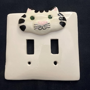 May include: White ceramic double light switch cover with a hand-painted cat face. The cat has black spots, green eyes, and a pink nose and mouth. The switch plate has two rectangular openings for the switches.