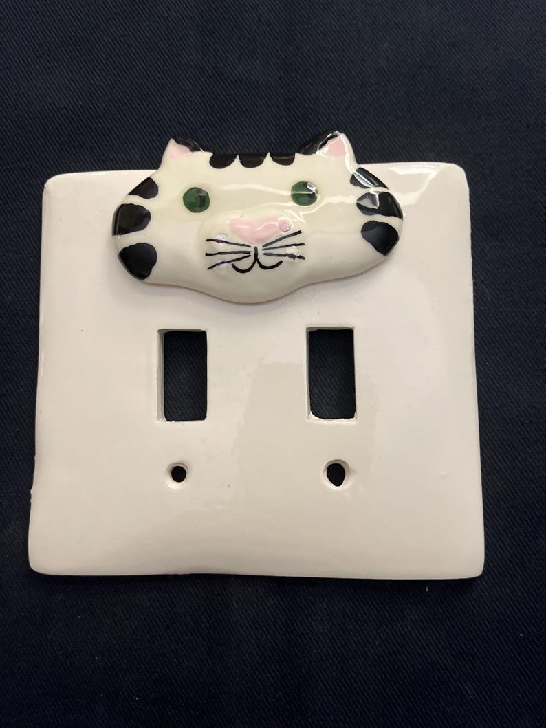 May include: White ceramic double light switch cover with a hand-painted cat face. The cat has black spots, green eyes, and a pink nose and mouth. The switch plate has two rectangular openings for the switches.