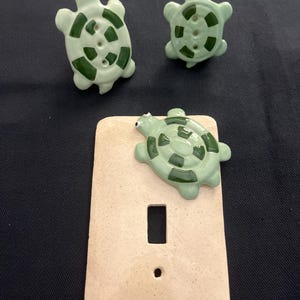 May include: A light switch cover with a green ceramic turtle design. The turtle is on the top of the cover and has a light green shell with dark green markings. The cover is a light beige color.