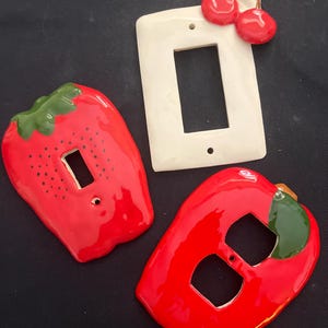 May include: Three ceramic light switch covers. One is white with a cherry design, and the other two are red, shaped like a strawberry and a pepper. The strawberry has black dots.