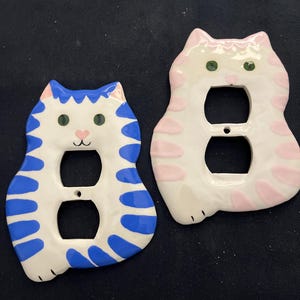 May include: Two ceramic cat-shaped light switch covers. One is white with blue stripes and accents, the other is white with pink stripes and accents. Both have green eyes and a pink nose. The covers are designed to fit standard electrical outlets.