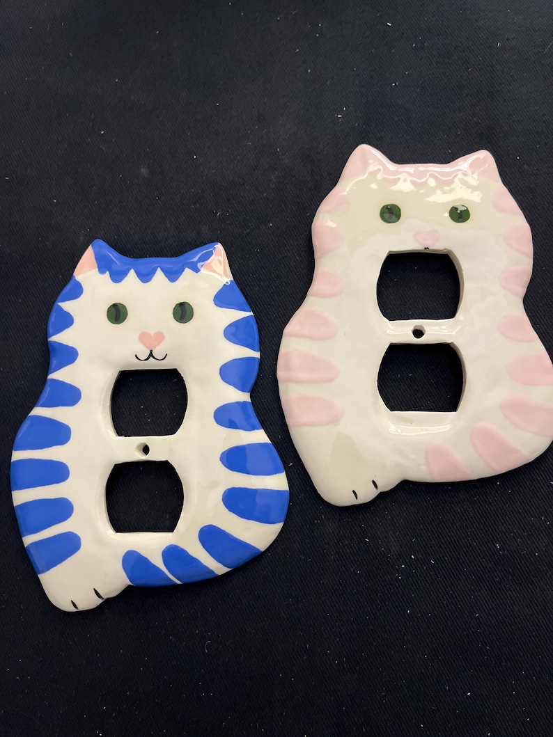 May include: Two ceramic cat-shaped light switch covers. One is white with blue stripes and accents, the other is white with pink stripes and accents. Both have green eyes and a pink nose. The covers are designed to fit standard electrical outlets.