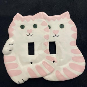 May include: A white ceramic double light switch cover featuring two cat shapes. Each cat has pink stripes, green eyes, and a pink heart-shaped nose. The cover has two rectangular openings for the light switches and two screw holes.