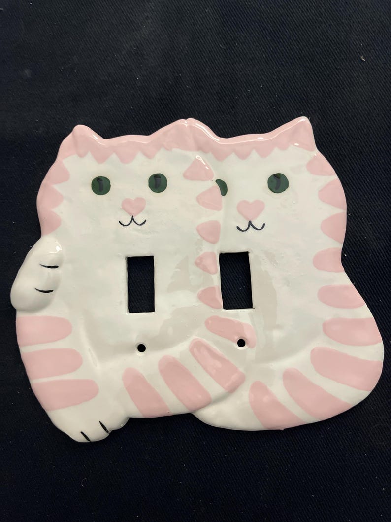 May include: A white ceramic double light switch cover featuring two cat shapes. Each cat has pink stripes, green eyes, and a pink heart-shaped nose. The cover has two rectangular openings for the light switches and two screw holes.