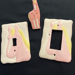 May include: Two ceramic light switch plates and one small horse figurine, all featuring a glossy finish with a hand-painted design of a pink horse with a yellow mane against a cream-colored background. The plates have rectangular cutouts.