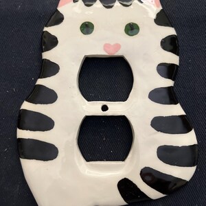 May include: A ceramic cat-shaped outlet cover. The cover is white with black stripes and spots, green eyes, and a pink nose and ears. The cover has two openings for electrical outlets.