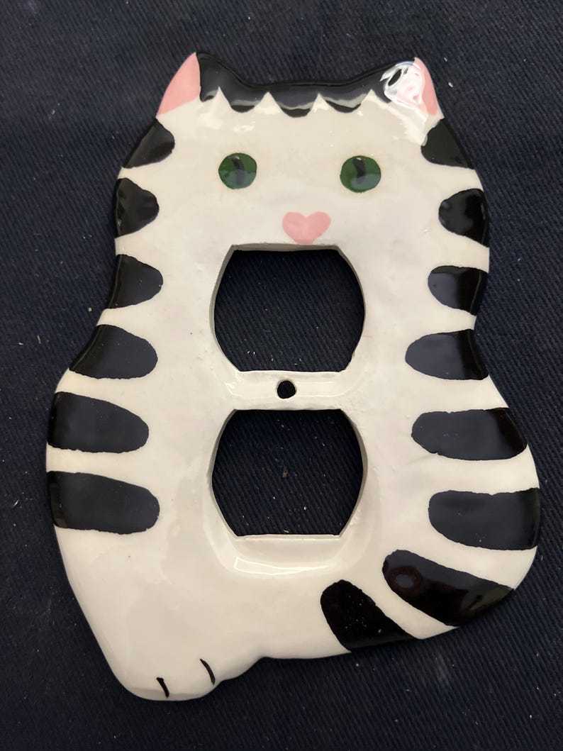 May include: A ceramic cat-shaped outlet cover. The cover is white with black stripes and spots, green eyes, and a pink nose and ears. The cover has two openings for electrical outlets.