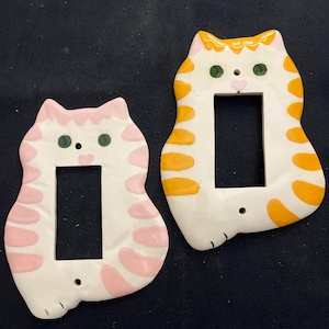 May include: Two ceramic cat-shaped light switch covers. One is white with pink accents, the other is white with orange stripes. Both have green eyes and a rectangular opening for the switch. The covers are designed to fit standard light switches.