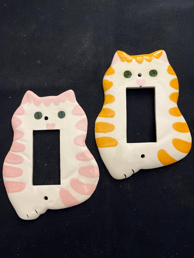 May include: Two ceramic cat-shaped light switch covers. One is white with pink accents, the other is white with orange stripes. Both have green eyes and a rectangular opening for the switch. The covers are designed to fit standard light switches.