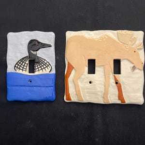 May include: Two ceramic light switch covers. One features a black and white loon over a blue and gray background. The other depicts a tan moose with brown legs and antlers on a cream-colored background. Both have rectangular cutouts.