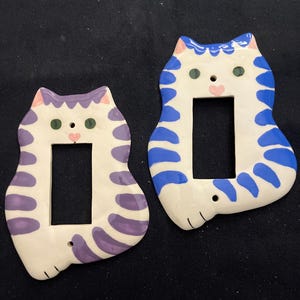 May include: Two ceramic light switch covers shaped like cats. One is white with purple stripes and pink ears, the other is white with blue stripes and blue ears. Both have green eyes and pink noses.