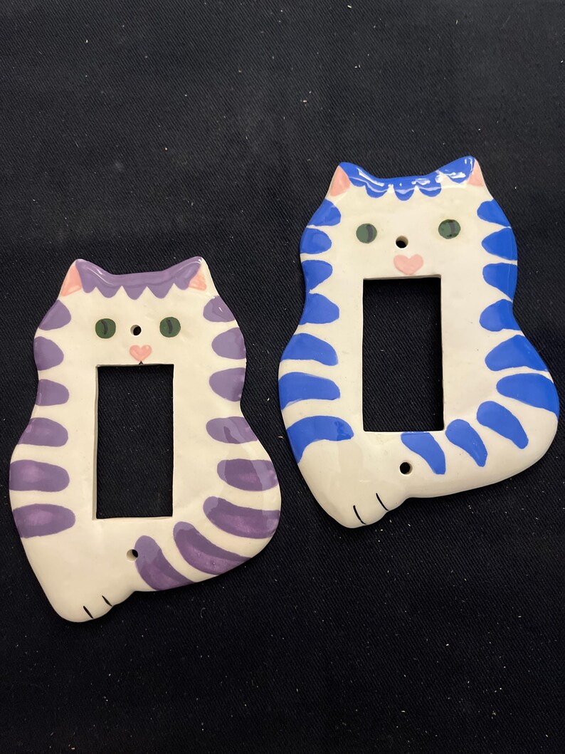 May include: Two ceramic light switch covers shaped like cats. One is white with purple stripes and pink ears, the other is white with blue stripes and blue ears. Both have green eyes and pink noses.