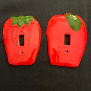 May include: Two ceramic light switch covers shaped like a strawberry and an apple. Both are red with green leaves and have a white light switch cutout.