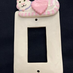 May include: A white ceramic light switch cover featuring a sleeping cat design. The cat has pink and white stripes, a pink heart, and a purple collar. The cover has a rectangular opening and two screw holes.