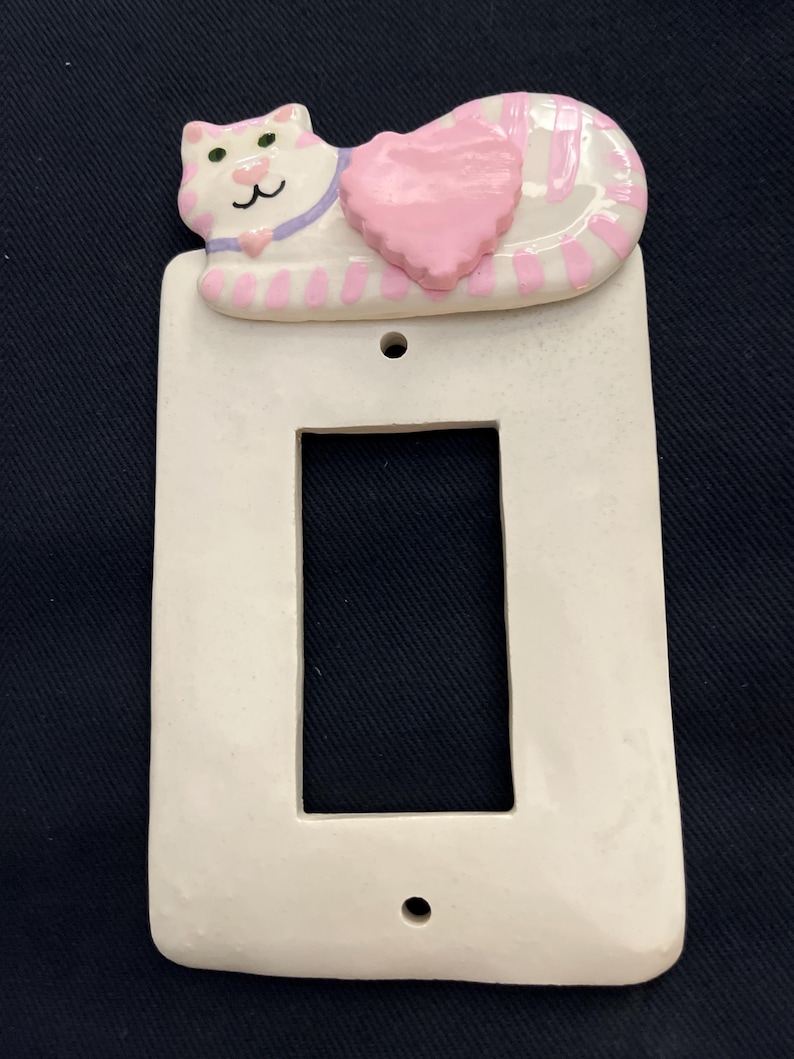 May include: A white ceramic light switch cover featuring a sleeping cat design. The cat has pink and white stripes, a pink heart, and a purple collar. The cover has a rectangular opening and two screw holes.