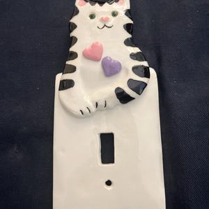 May include: A ceramic light switch cover featuring a white cat with black markings and pink accents. The cat has two heart-shaped embellishments, one pink and one purple. The switch plate is white with a rectangular opening for the switch.