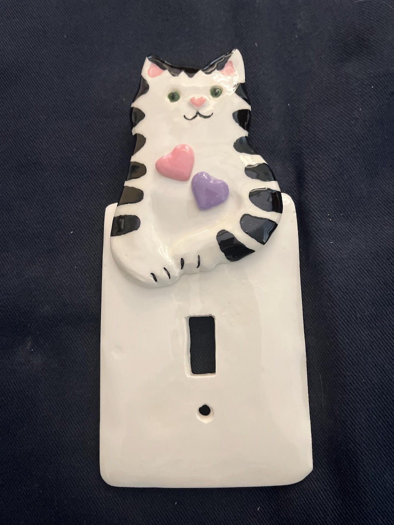 May include: A ceramic light switch cover featuring a white cat with black markings and pink accents. The cat has two heart-shaped embellishments, one pink and one purple. The switch plate is white with a rectangular opening for the switch.